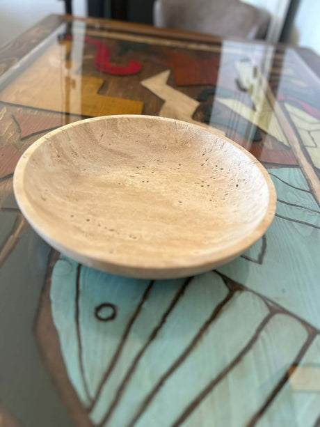 The Timeless Travertine Artisanal Bowl by Travertable, with its smooth finish and visible grain patterns, rests on a glass tabletop showcasing a colorful abstract design, bringing elegance and creativity to your space.