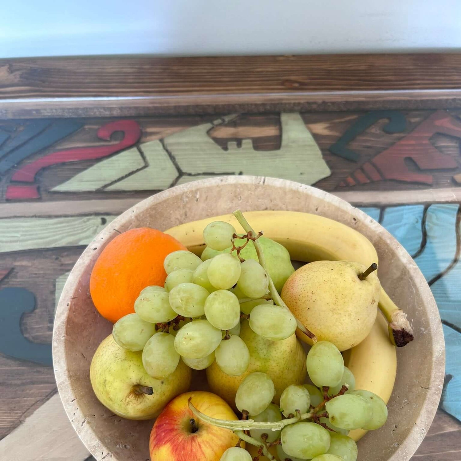 The Timeless Travertine Artisanal Bowl by Travertable holds green grapes, a banana, an orange, an apple, and two pears on a vibrant table with abstract designs.