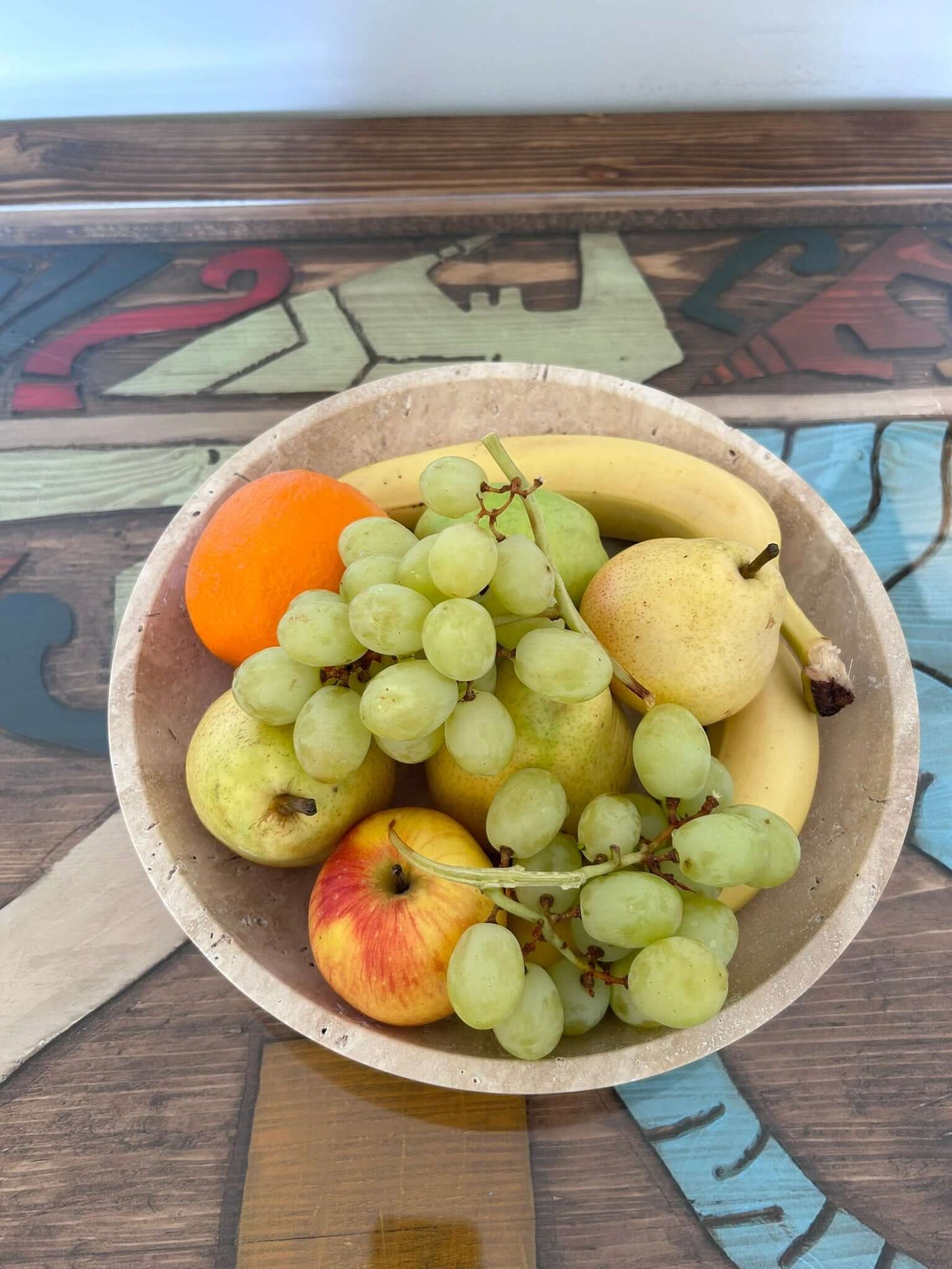 The Timeless Travertine Artisanal Bowl by Travertable holds green grapes, a banana, an orange, an apple, and two pears on a vibrant table with abstract designs.