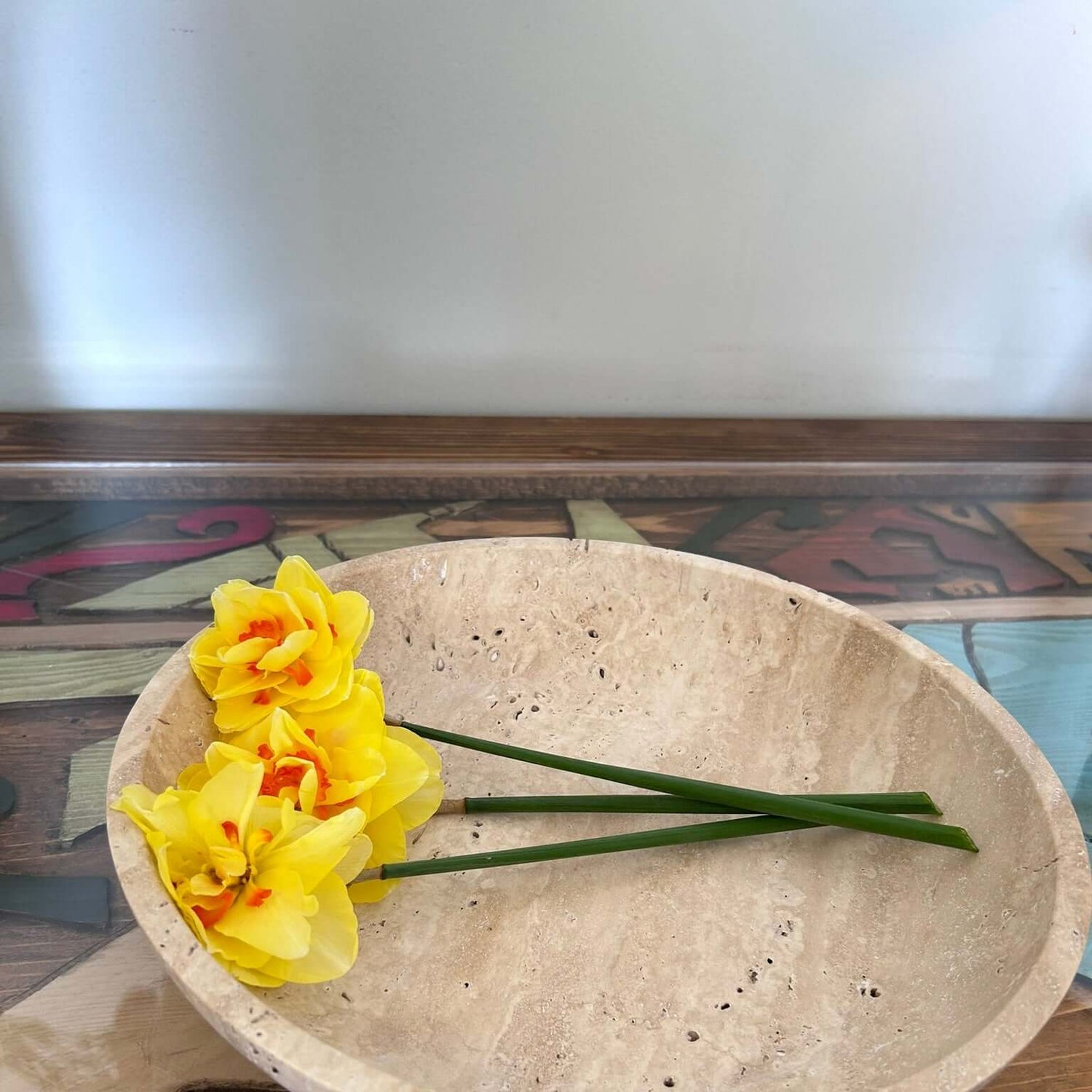 A Timeless Travertine Artisanal Bowl from Travertable sits on a colorful wooden table. Inside, three yellow daffodils with orange centers lie flat, showcasing natural artistry. The slightly blurred background highlights the bowl and flowers.