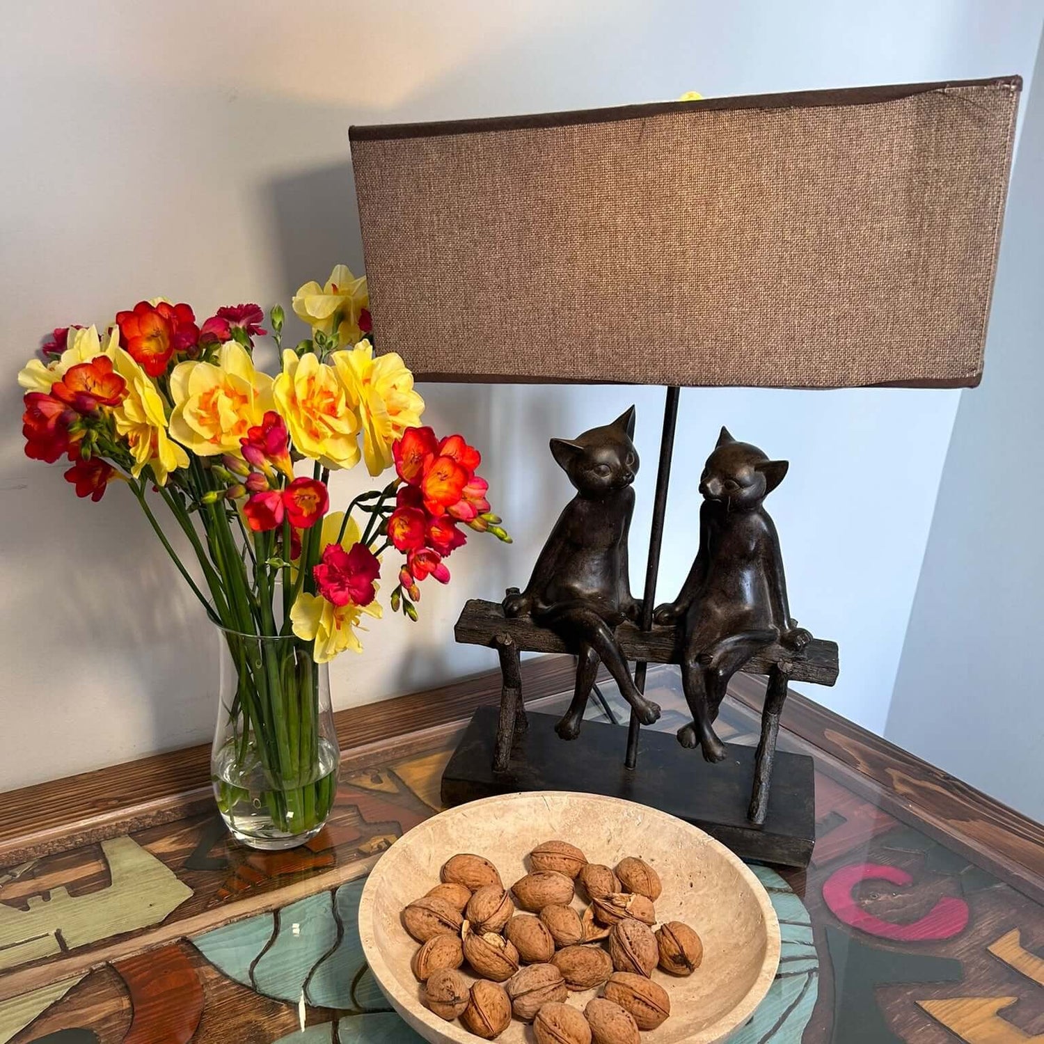 A Timeless Travertine Artisanal Bowl by Travertable, filled with unshelled almonds, sits on a glass-top table with abstract art beneath. A vase of red and yellow flowers and a sculpted lamp with two sitting cats add to the natural artistry of the scene.