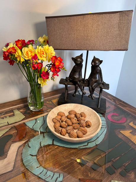 A Timeless Travertine Artisanal Bowl by Travertable, filled with unshelled almonds, sits on a glass-top table with abstract art beneath. A vase of red and yellow flowers and a sculpted lamp with two sitting cats add to the natural artistry of the scene.