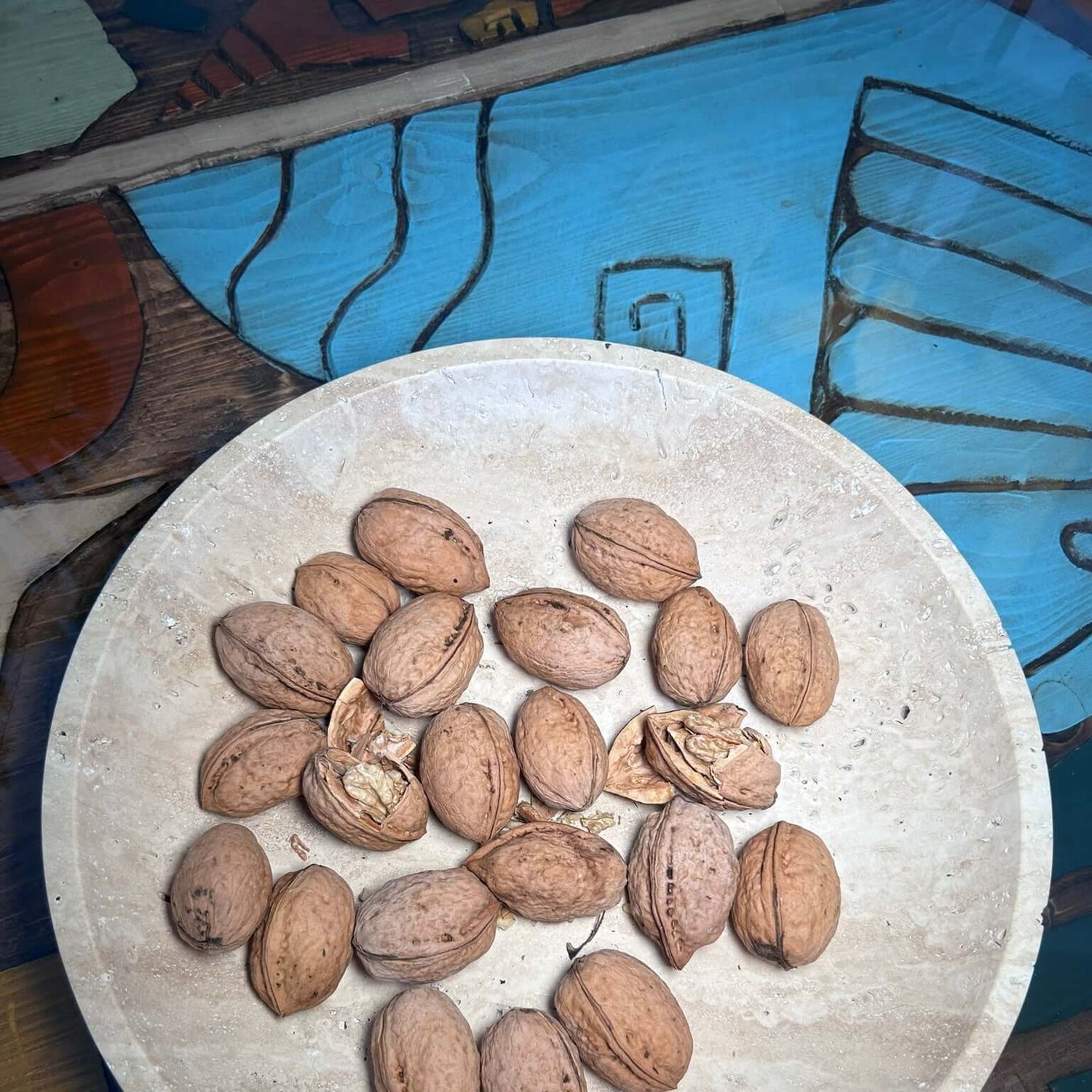 The Timeless Travertine Artisanal Bowl by Travertable elegantly holds walnuts. It rests on a vibrant, patterned surface in hues of blue, brown, and teal, accentuating its natural artistry.
