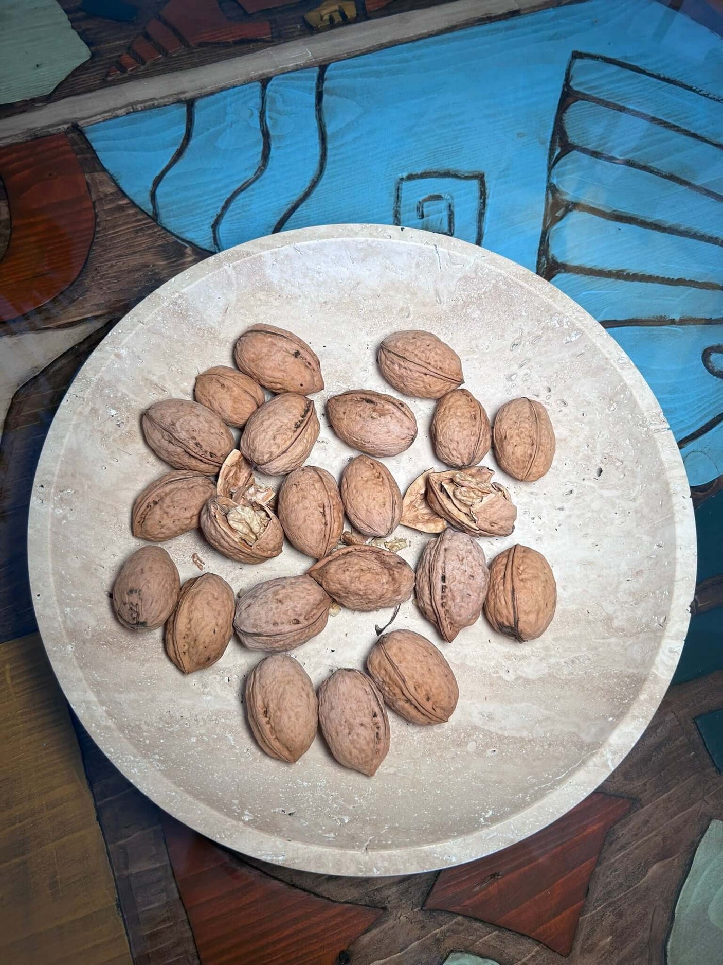 The Timeless Travertine Artisanal Bowl by Travertable elegantly holds walnuts. It rests on a vibrant, patterned surface in hues of blue, brown, and teal, accentuating its natural artistry.
