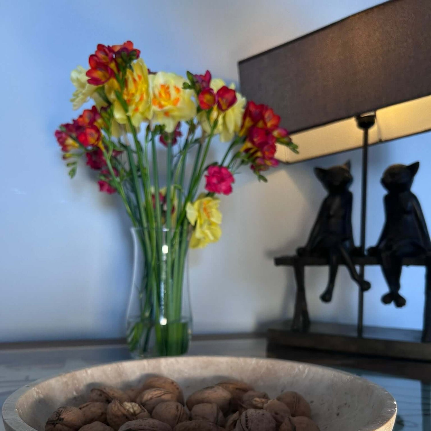 A Timeless Travertine Artisanal Bowl from Travertable sits on a glass table. Behind it, a vase with yellow and red flowers complements a lamp adorned with two cat figurines, adding to the rooms natural artistry.