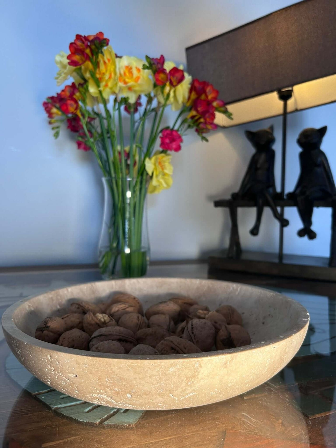 A Timeless Travertine Artisanal Bowl from Travertable sits on a glass table. Behind it, a vase with yellow and red flowers complements a lamp adorned with two cat figurines, adding to the rooms natural artistry.