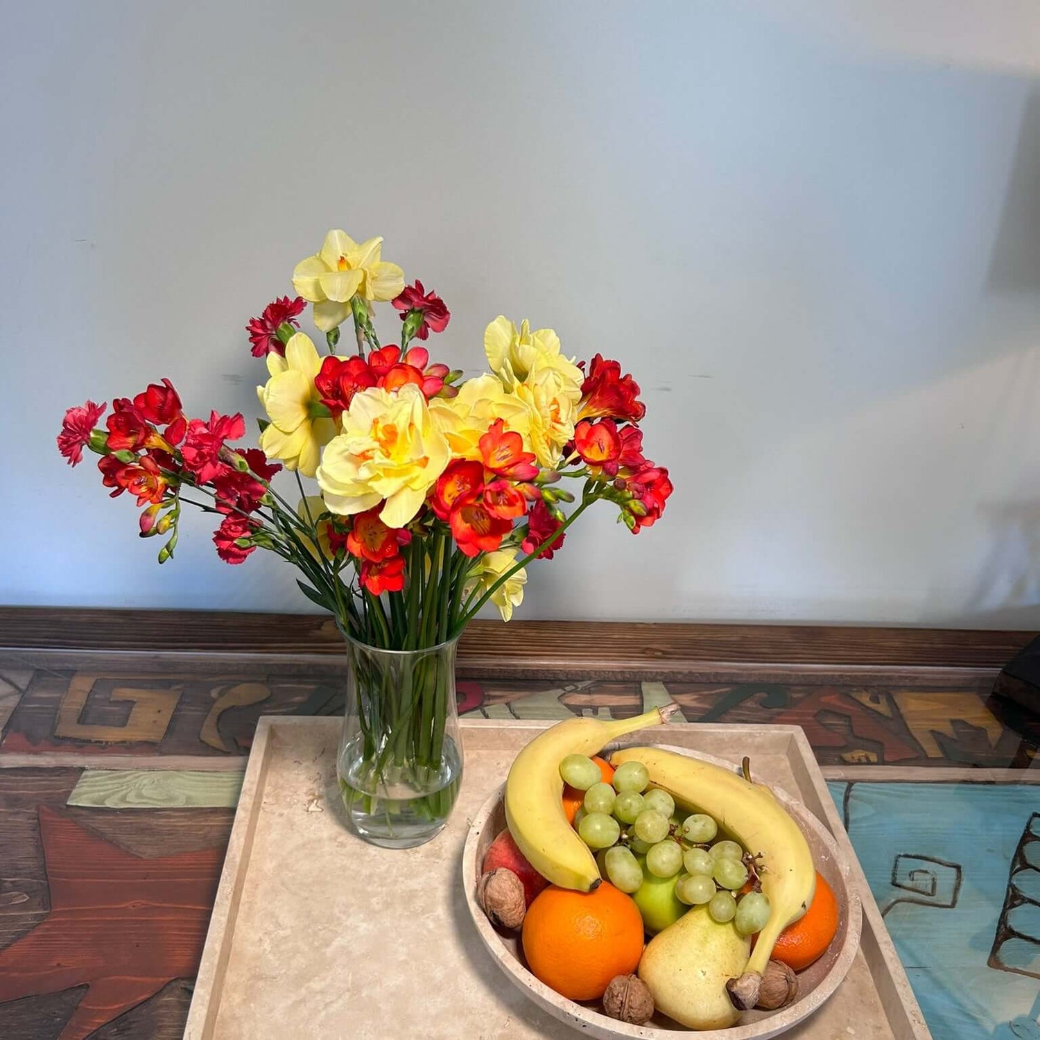 A vase with red and yellow flowers complements a Timeless Travertine Artisanal Bowl by Travertable filled with bananas, green grapes, oranges, pears, and walnuts on a tray. This ensemble sits on a colorful wooden surface that highlights natural artistry in every detail.