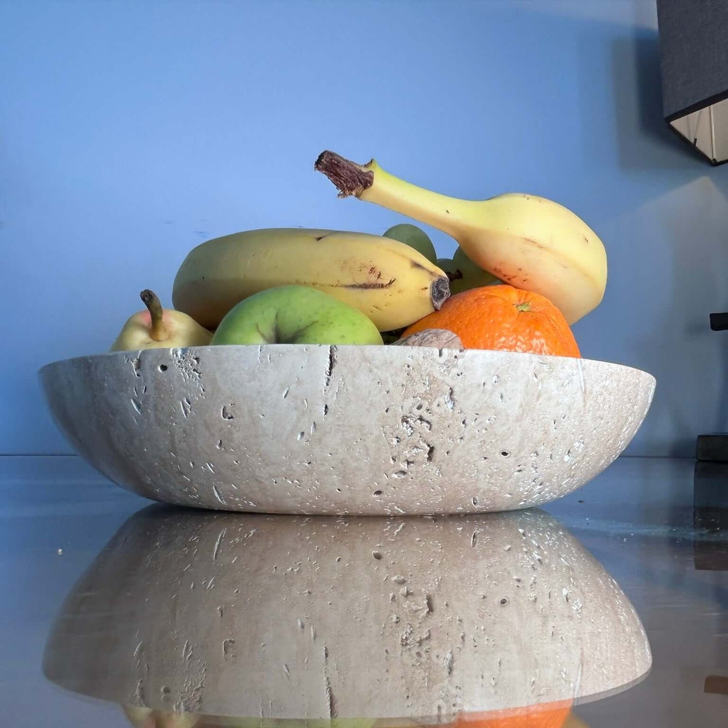 The Timeless Travertine Artisanal Bowl by Travertable holds bananas, an apple, a pear, and an orange on a shiny table reflecting a smooth blue wall.