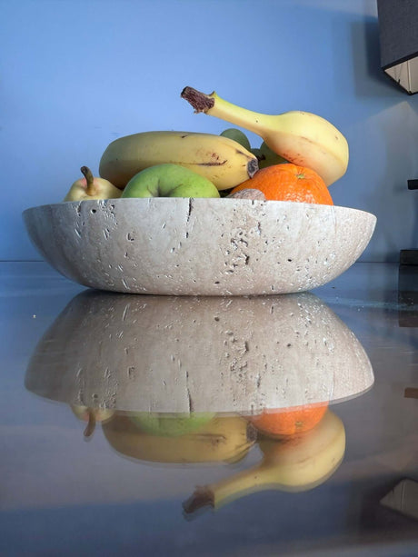 The Timeless Travertine Artisanal Bowl by Travertable holds bananas, an apple, a pear, and an orange on a shiny table reflecting a smooth blue wall.