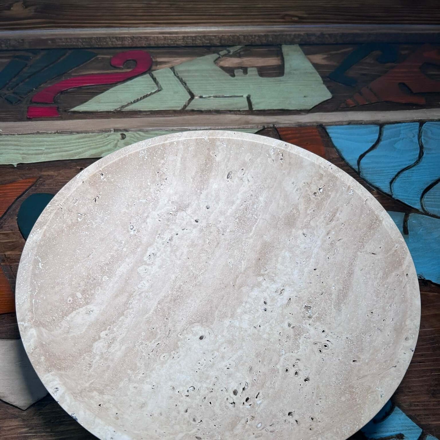 A round, beige marble bowl with natural veining and small pockmarks rests on a wooden surface featuring abstract designs. The Timeless Travertine Artisanal Bowl by Travertable complements the table, spotlighting the harmony of smooth texture and natural artistry.