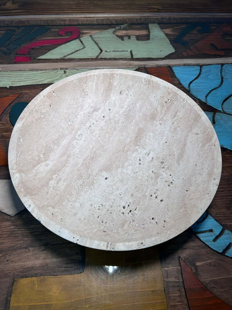 A round, beige marble bowl with natural veining and small pockmarks rests on a wooden surface featuring abstract designs. The Timeless Travertine Artisanal Bowl by Travertable complements the table, spotlighting the harmony of smooth texture and natural artistry.