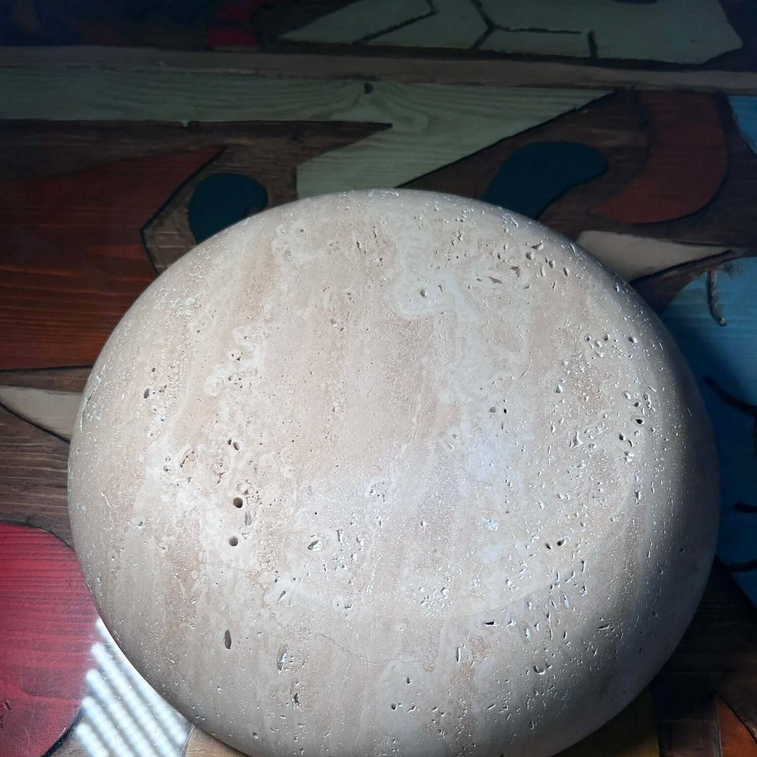 The Timeless Travertine Artisanal Bowl by Travertable features a round, textured stone surface with small holes, and is set against a colorful, patterned background.