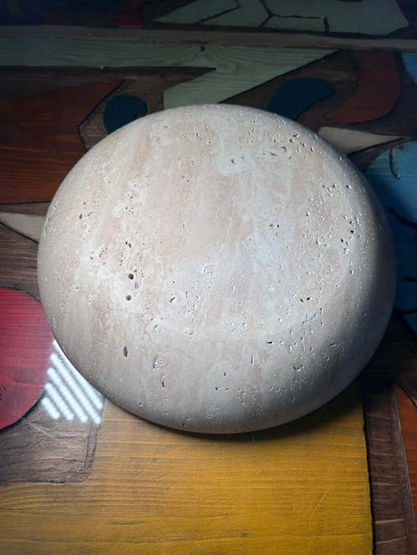 The Timeless Travertine Artisanal Bowl by Travertable features a round, textured stone surface with small holes, and is set against a colorful, patterned background.