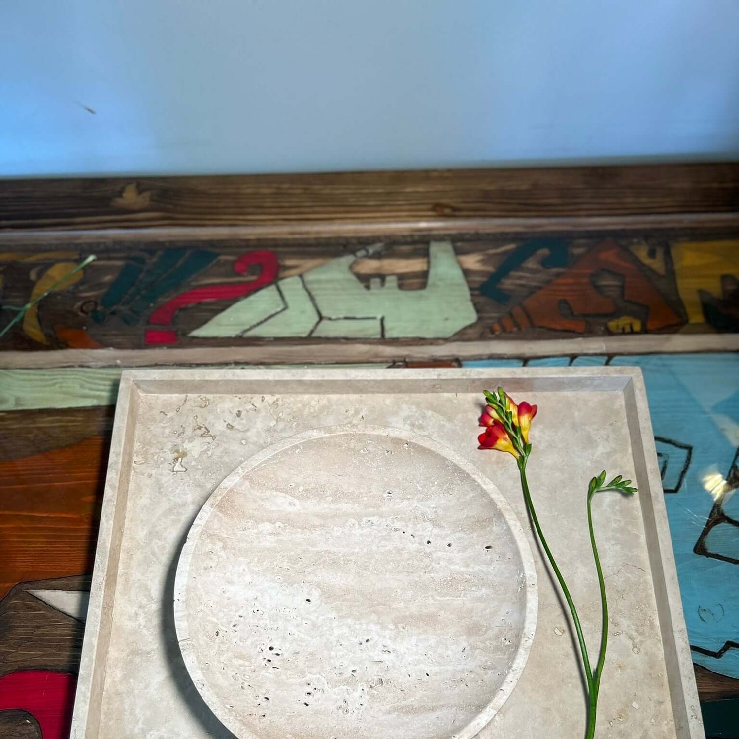 The Timeless Travertine Artisanal Bowl by Travertable is centered on a square patterned stone slab, with a red and yellow flower and green stems beside it against a multicolored abstract backdrop, showcasing natural artistry in its simplest form.