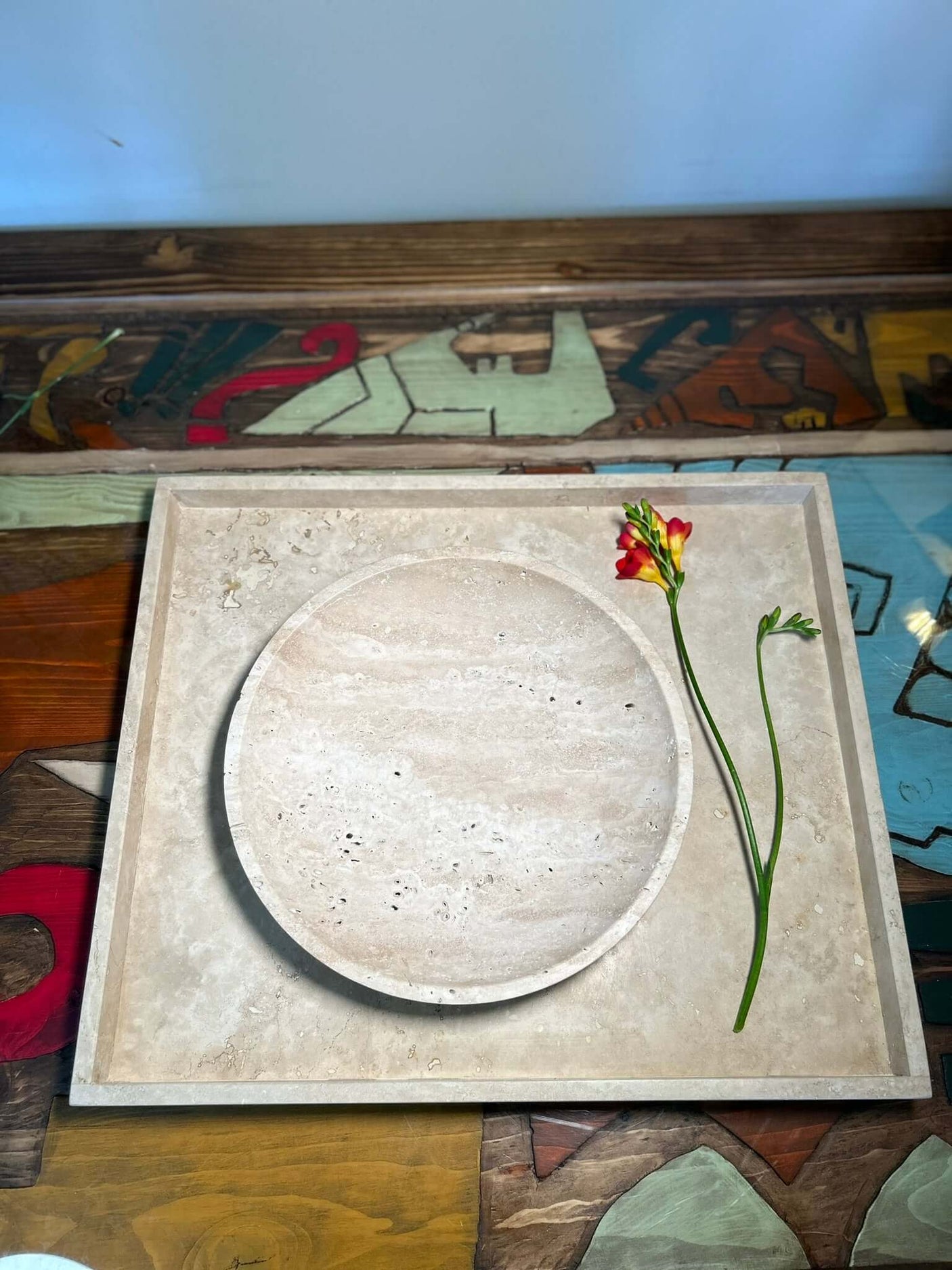 The Timeless Travertine Artisanal Bowl by Travertable is centered on a square patterned stone slab, with a red and yellow flower and green stems beside it against a multicolored abstract backdrop, showcasing natural artistry in its simplest form.