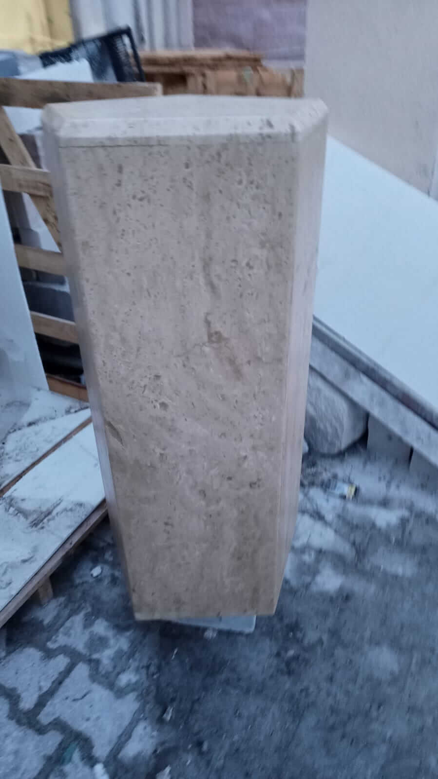 A Geometric Travertine Pedestal by Travertable stands upright on a paved surface, surrounded by wooden pallets and construction materials. With its textured surface and uneven top edge, this pedestal echoes the artistry of sculptural columns in an unfinished form.