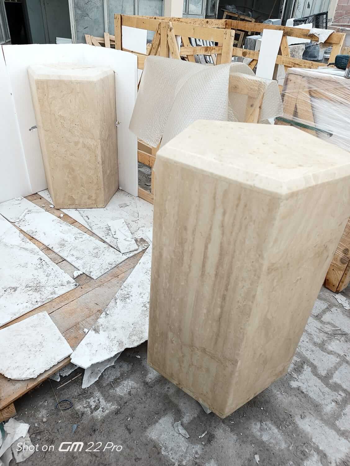 Among construction materials like wooden crates and tiles, two large, hexagonal Geometric Travertine Pedestals from Travertable stand out with their unique sculptural design and light travertine resemblance in an unfinished setting.