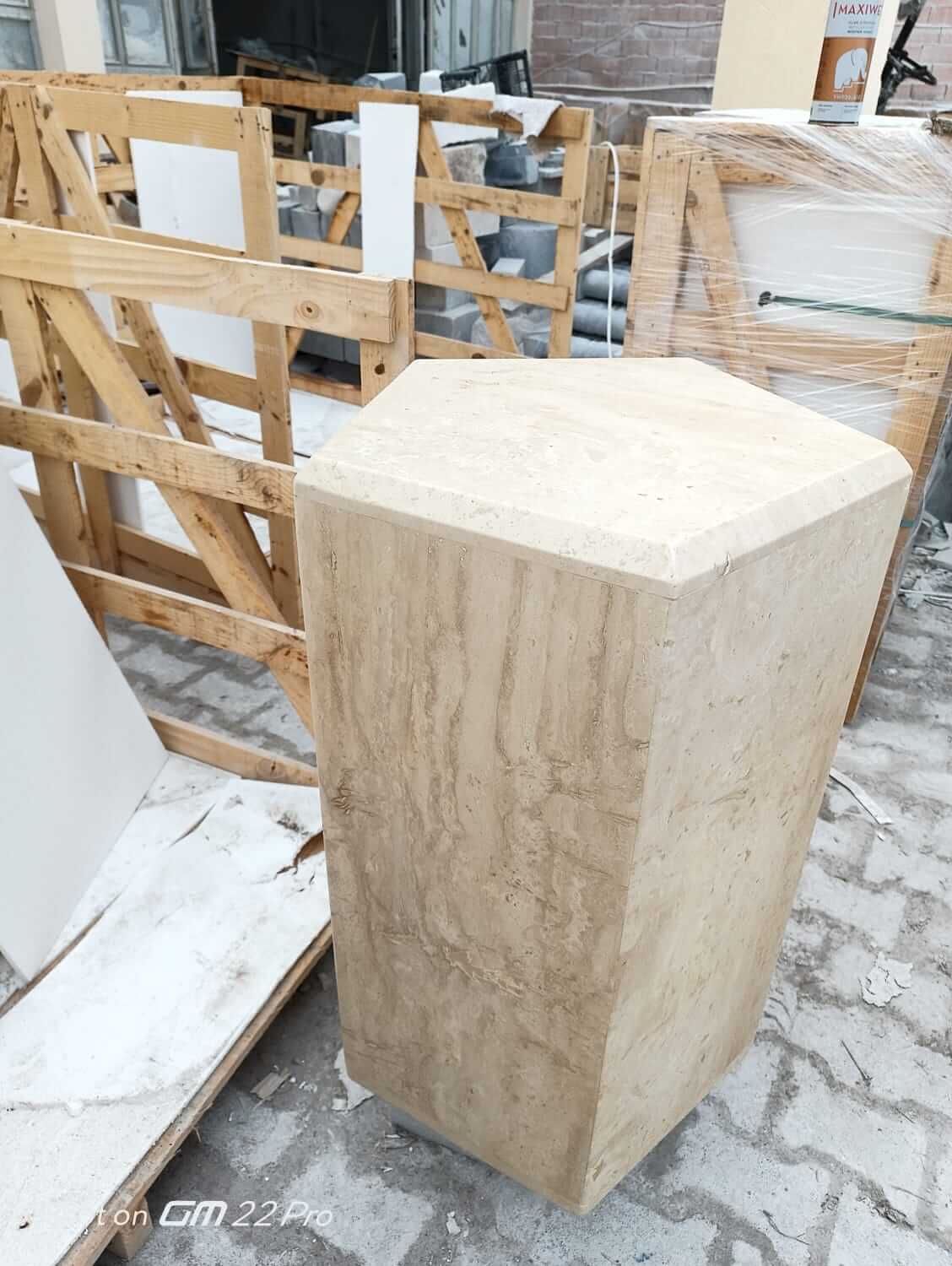 The Geometric Travertine Pedestal by Travertable, featuring a unique sculptural design with architectural appeal, stands upright on a workshop floor. Behind it lie scattered wooden frames and materials, creating an industrial or construction-like setting.