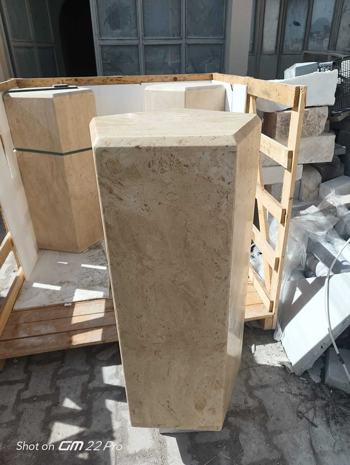 The Geometric Travertine Pedestal by Travertable, with its unique sculptural design, stands outdoors on a wooden pallet. Made from light travertine, it exhibits a light brown hue and natural patterns that catch sunlight, casting shadows across the ground amidst other stone slabs and materials.