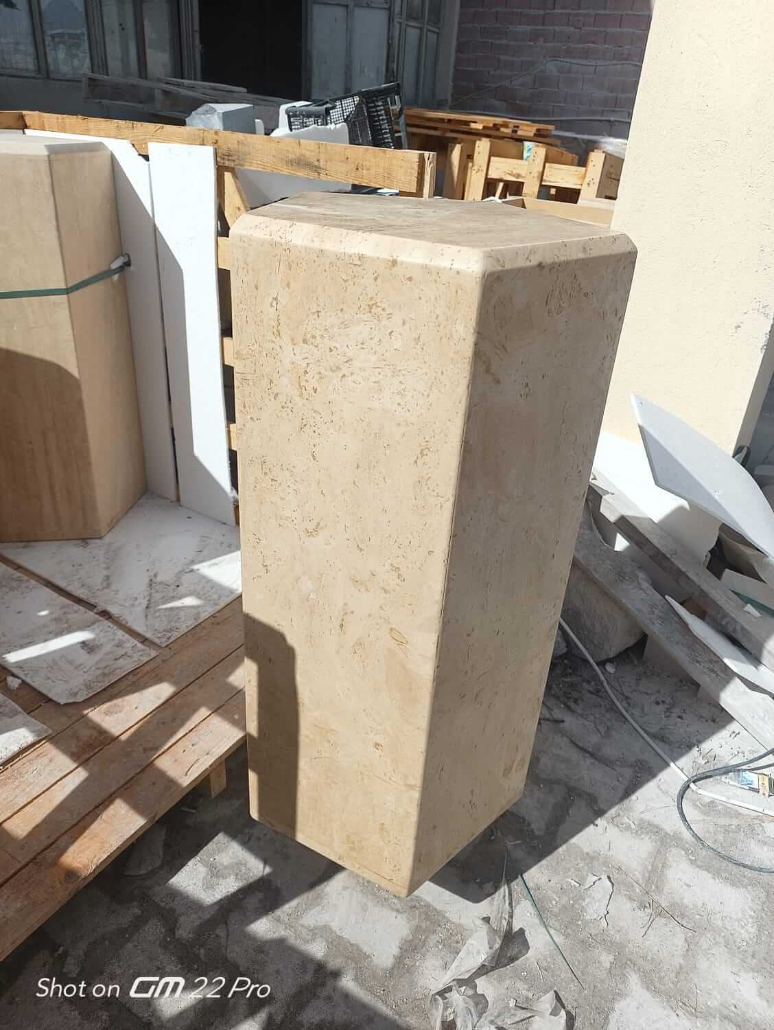 The Geometric Travertine Pedestal by Travertable, a large rectangular stone block with a smooth beige surface and sculptural charm, stands upright on pavement surrounded by wooden pallets. Sunlight casts shadows, enhancing its architectural appeal.