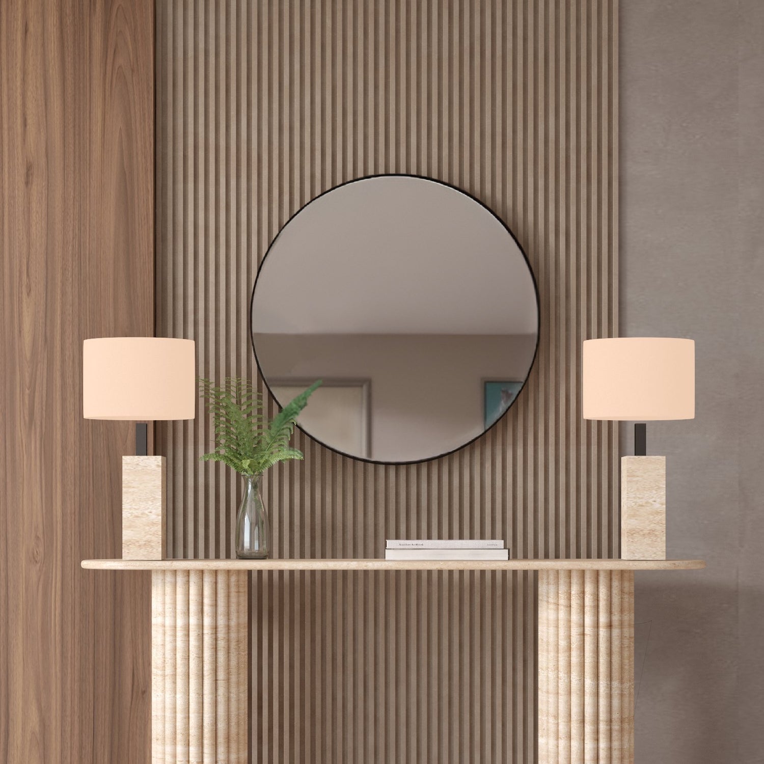 The Ovale Travertine Console Table by TRAVERTABLE features a vertical groove organic stone base and is customizable. Styled here with beige lamps, a glass vase with green leaves, and a round mirror on a wood slat accent wall behind it.