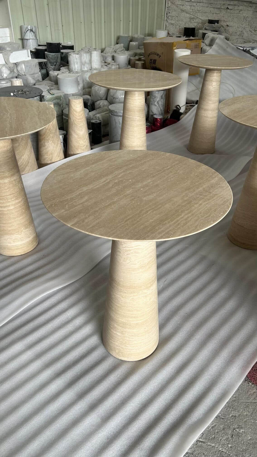 The Twin Pillars Travertine Side Table Set by Travertable, with its natural stone elegance and conical bases, stands in an industrial setting on protective white material, surrounded by assorted packaging items.