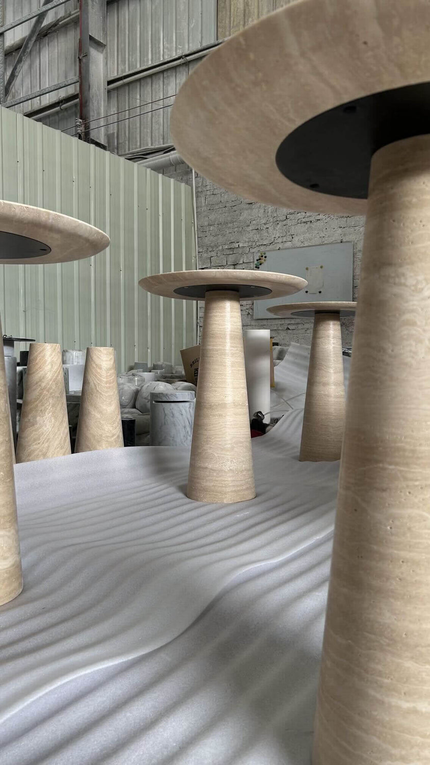 Travertables Twin Pillars Travertine Side Table Set, featuring round beige tables with cone-shaped bases, is displayed upside down on a white textured surface in a warehouse. The industrial space includes corrugated metal walls and concrete bricks, exuding natural elegance.