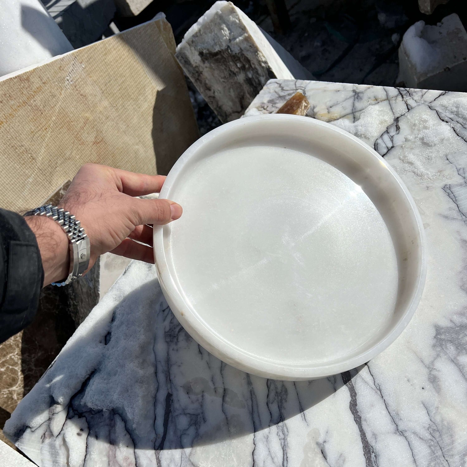 A hand presents the Travertable White Alabaster Round Tray over a marble surface with gray veining. The outdoor setting and the polished tray elevate luxury decor, enhancing a sophisticated ambiance.