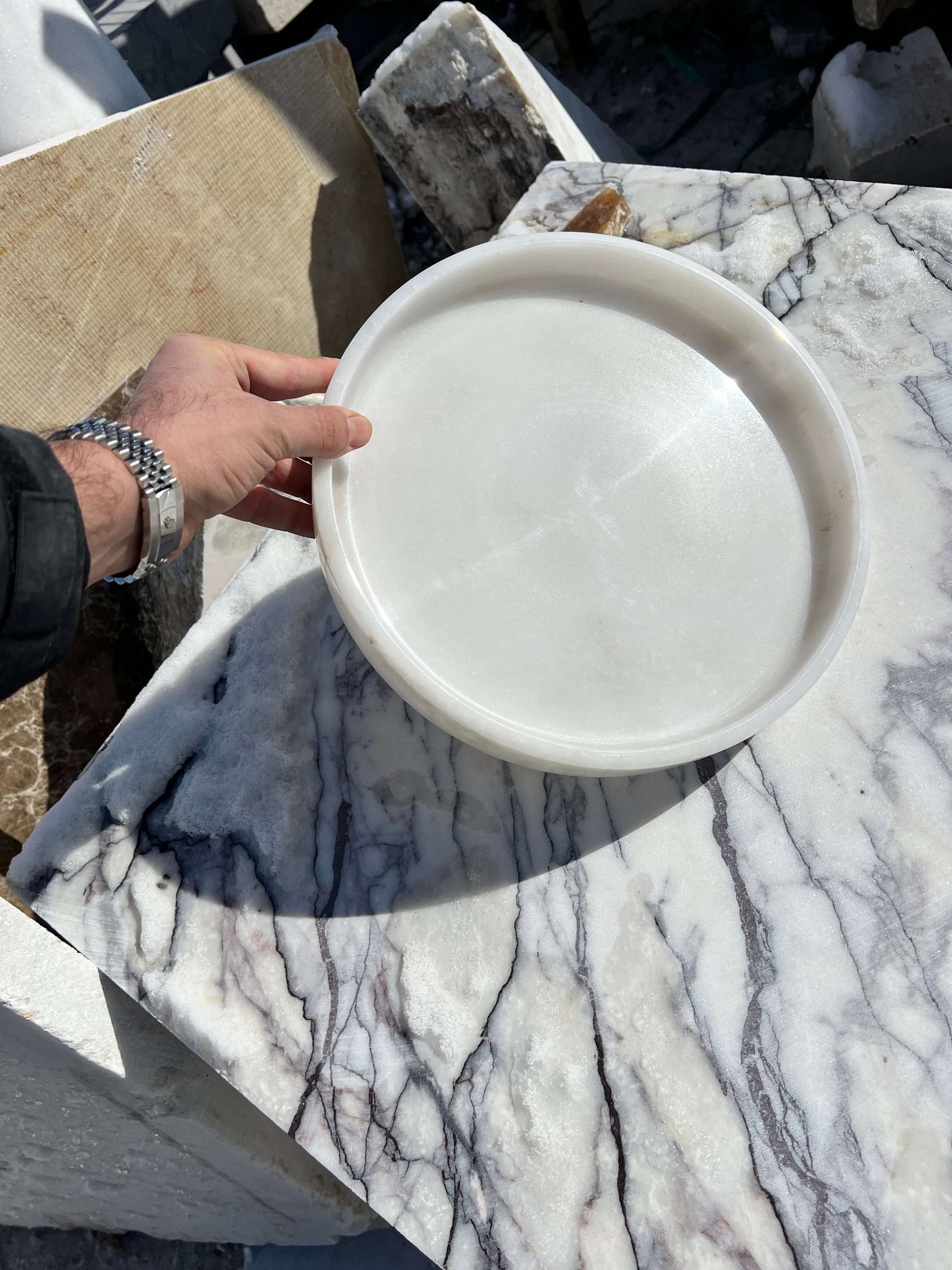 A hand presents the Travertable White Alabaster Round Tray over a marble surface with gray veining. The outdoor setting and the polished tray elevate luxury decor, enhancing a sophisticated ambiance.