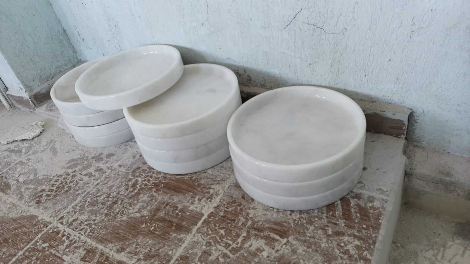 Travertables White Alabaster Round Trays, known as the Luxe Marble Accent Piece, are stacked on a dusty floor against a textured wall, bringing an elegant touch to home decor with their smooth and luxurious design.