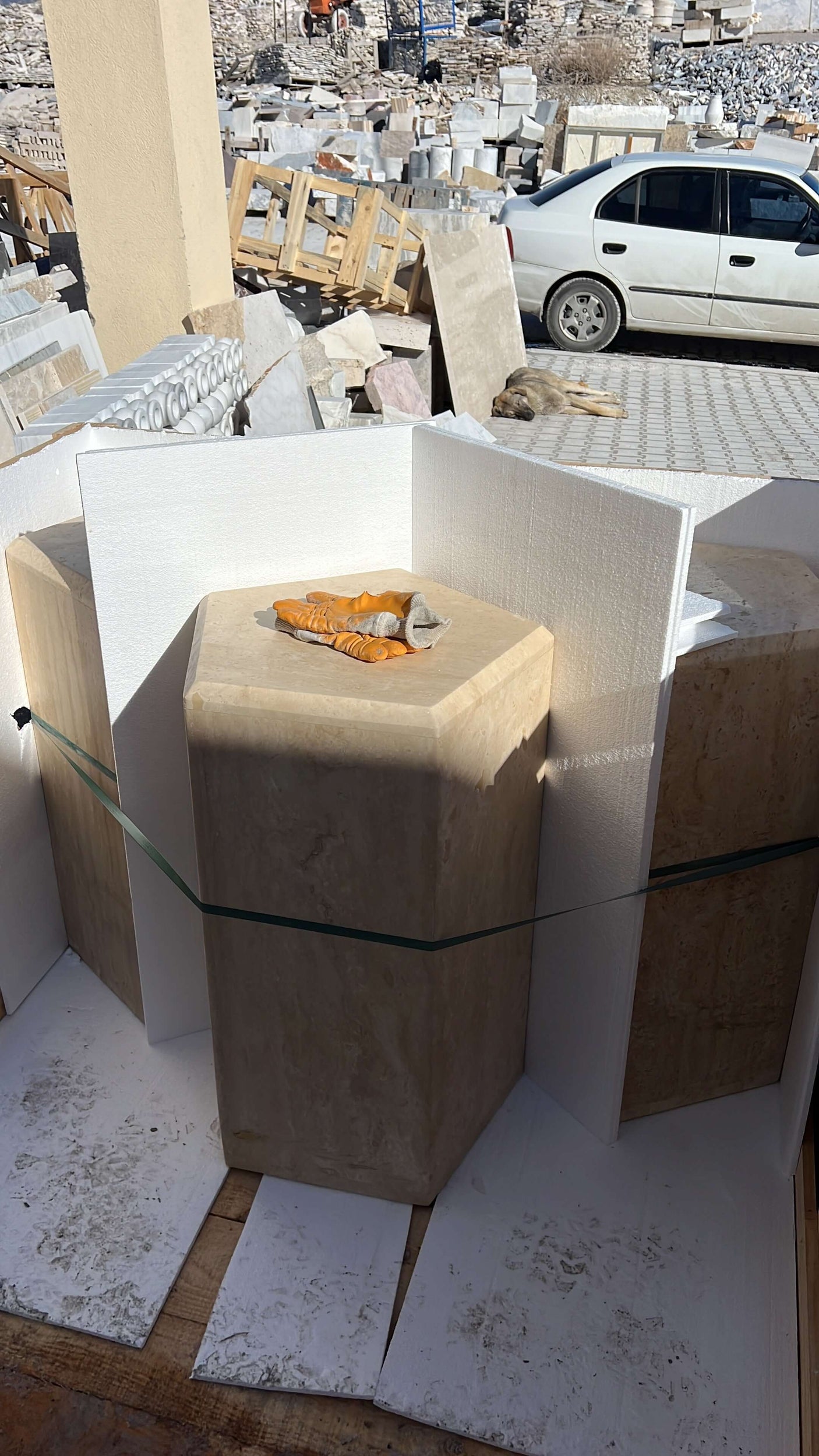 A set of large, beige Travertable Geometric Travertine Pedestals with unique sculptural design and architectural appeal stands neatly upright, surrounded by white protective panels. A chisel and wooden mallet rest on one pedestal. In the background, a white car is parked.