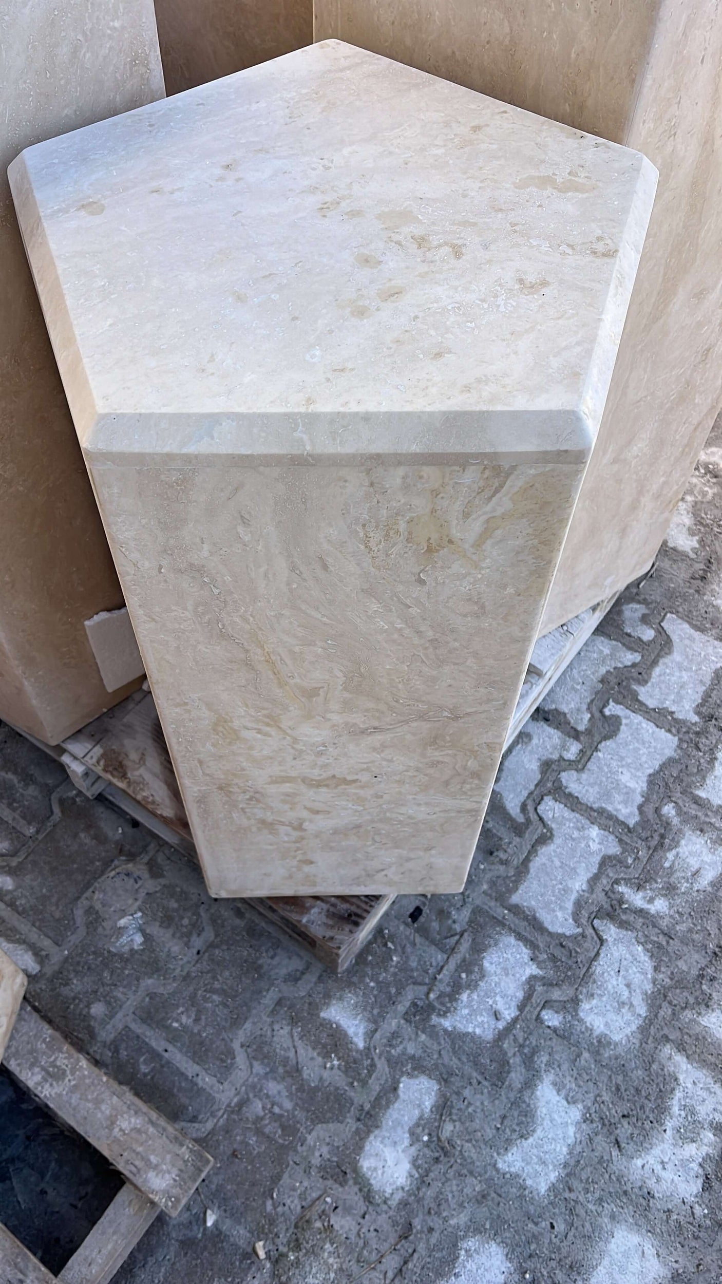 The Geometric Travertine Pedestal by Travertable features a polished hexagonal marble top with subtle natural patterns and variations. Its smooth, beige surface stands on a stone base, resembling light travertine pedestals. It is partially set on pallet wood for added design contrast.