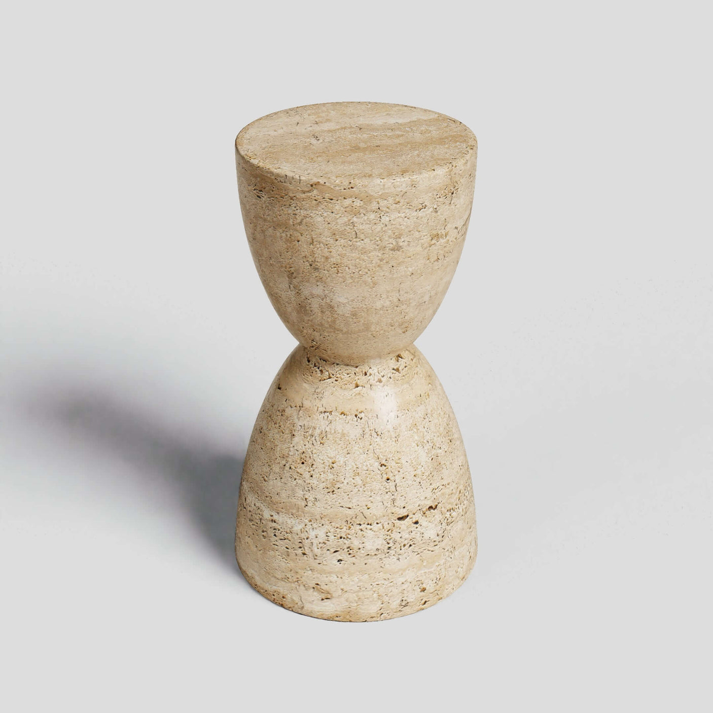 The Eda Beige Travertine Sphere Table by Travertable is a stone, hourglass-shaped stool with a rough texture, embodying minimalist design. Its symmetrical halves unite at the center, forming an abstract sculpture. Positioned against a plain gray backdrop, it casts a delicate shadow.