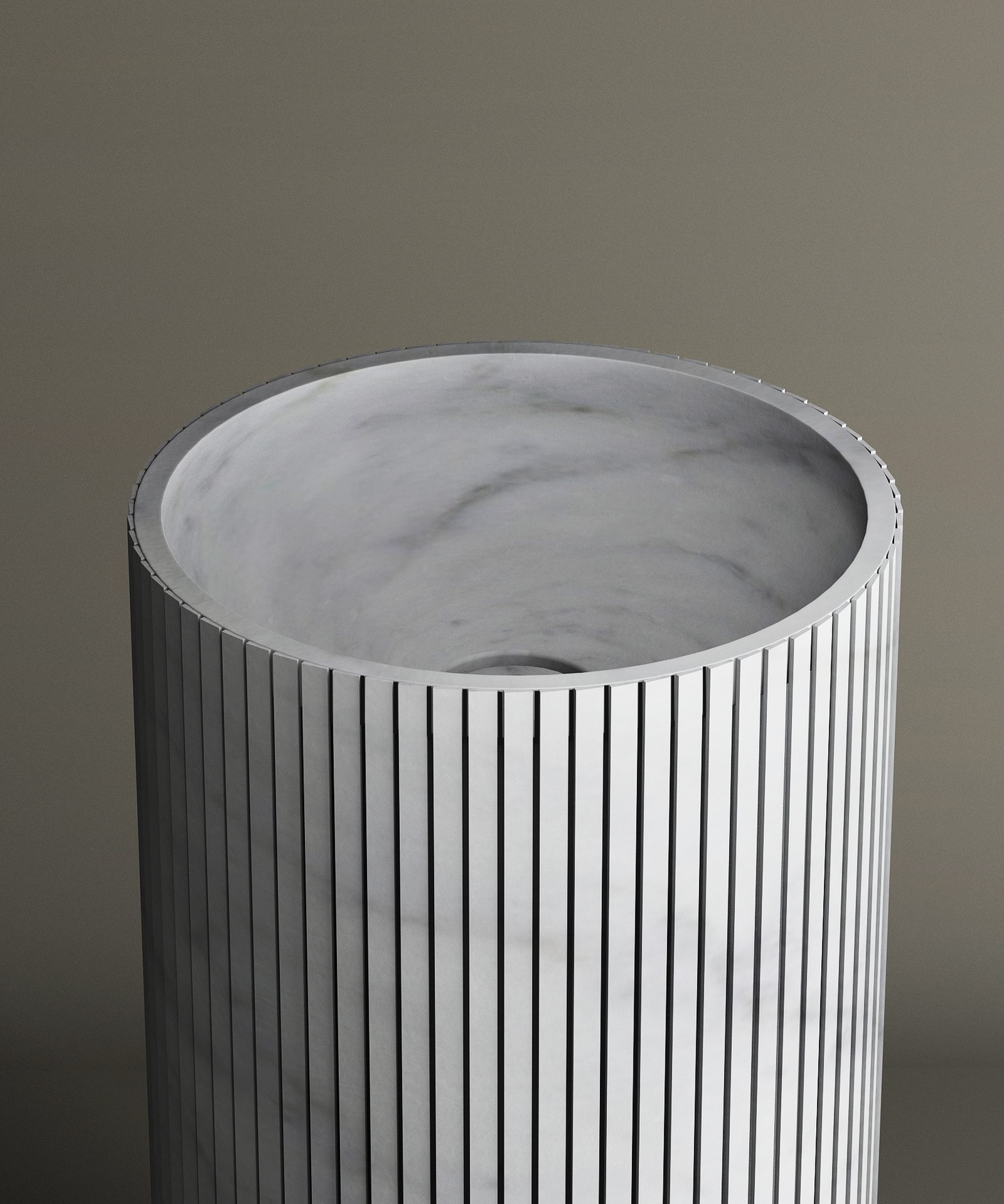 The TRAVERTABLE Carrara Round Fluted Marble Pedestal Vanity Sink features a cylindrical, fluted design with a marble texture and an open top revealing a smooth light gray interior, all set against a neutral background.