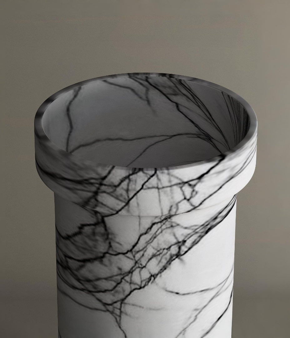 The TRAVERTABLE Lilac Marble Pedestal Vanity Sink features a freestanding cylindrical design with black, grey, and subtle lilac veining, an open top with a thick rim, and is shown against a plain neutral background.