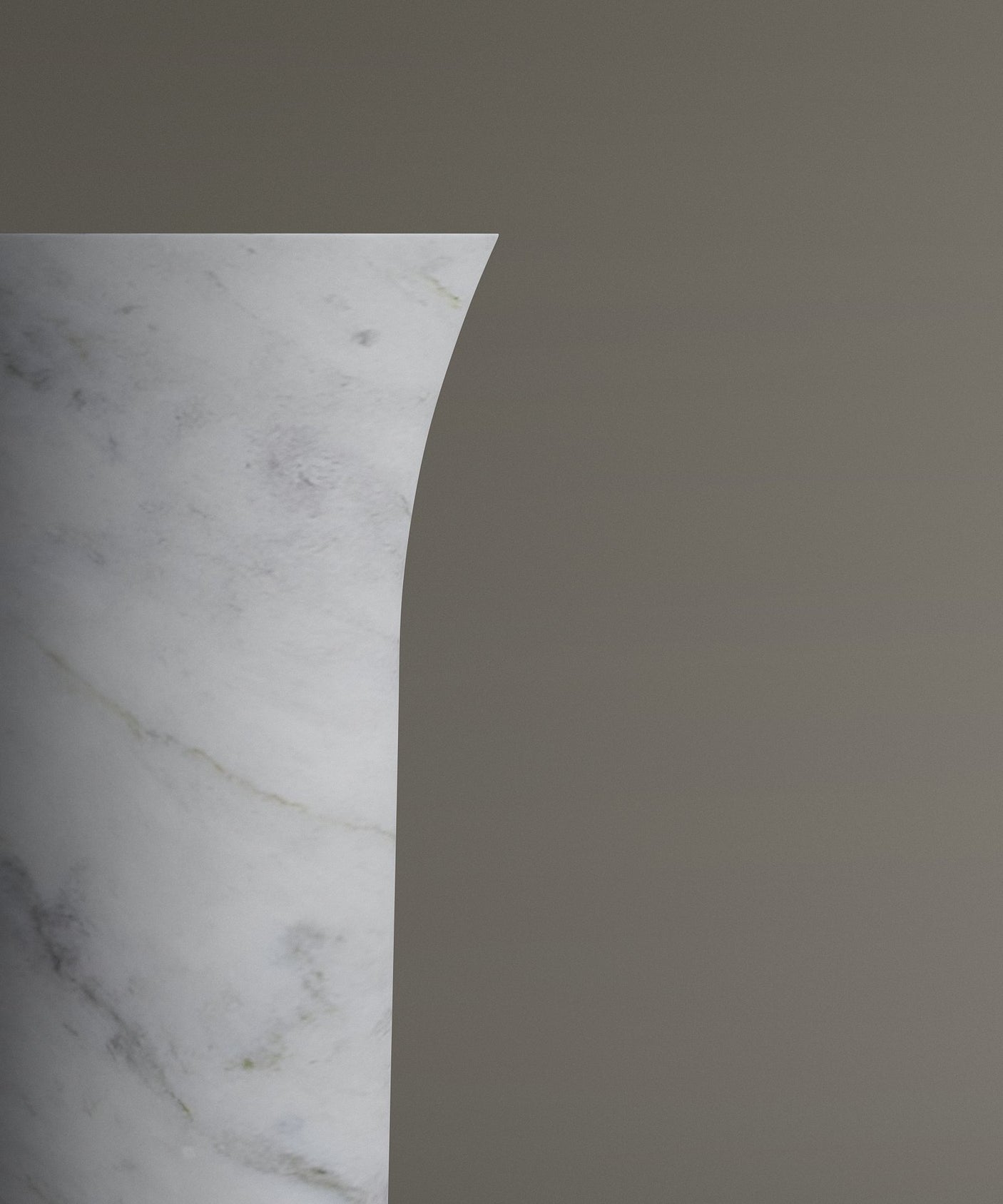 A close-up of the TRAVERTABLE Carrara Marble Cylinder Pedestal Vanity Sink shows its white surface with subtle gray veining, smooth texture, and curved edge, set against a plain taupe background.