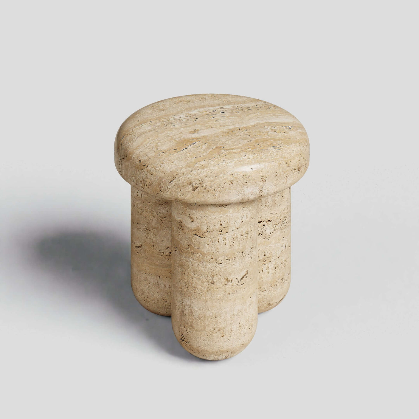 The Travertable Travertine Elegance Accent Table is a small, round end table made from natural stone with a light beige tone. It features a smooth top and three cylindrical legs, set on a plain gray background.