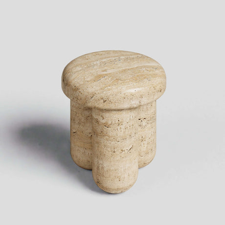 The Travertable Travertine Elegance Accent Table is a small, round end table made from natural stone with a light beige tone. It features a smooth top and three cylindrical legs, set on a plain gray background.