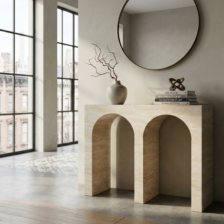 A modern entryway with a beige stone console table featuring three arches, two matching lamps with beige shades, a glass vase with green fern leaves, books, and a round mirror on the wall.