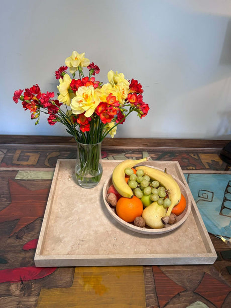A round, textured beige stone bowl and tray set sit on a colorful, abstract wooden table. A single stem with small red and yellow flowers rests on the tray beside the bowl.