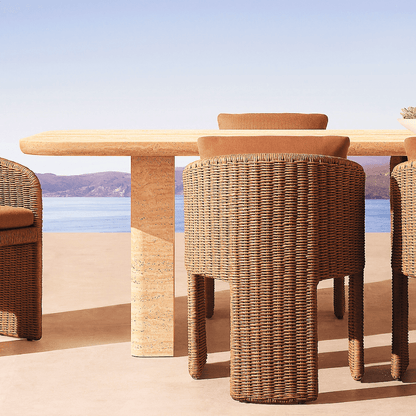 Brown-cushioned wicker chairs encircle The Majestic Travertine Dining Table by Travertable, providing a modern aesthetic. With customizable tabletop thickness, this patio setup overlooks a clear blue lake and distant hills under a perfect sky.