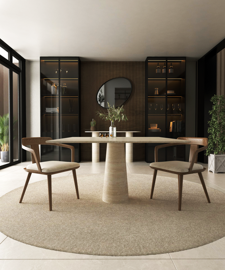 A modern dining room featuring the TRAVERTABLE Square Travertine Dining Table with Conical Pedestal Base, two wooden chairs, a circular rug, built-in shelves with decor, a round mirror on a wood-paneled wall, large windows, and green plants.