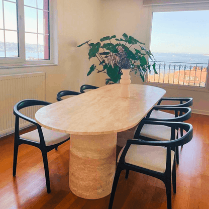 The dining room features an Arcadia Dining Table by Travertable, showcasing an elegant oval design. Surrounded by six black chairs with white cushions, a white vase with green plants adds charm. Large windows offer stunning sea views and distant hills, while a warm wooden floor enhances elegance.