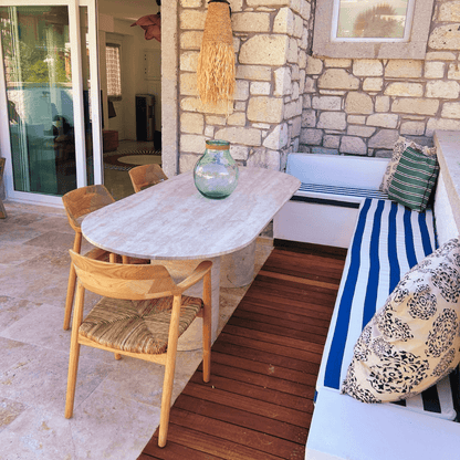 The outdoor patio, framed by a stone wall, features an Arcadia Dining Table by Travertable, surrounded by wicker chairs and a cushioned blue-and-white striped bench with patterned pillows. A large glass vase adorns the travertine surface as natural light filters through the glass doors.