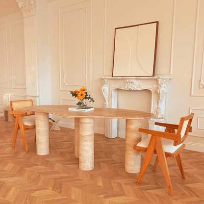 A chic interior with the Travertable Cascade Travertine Table featuring customizable options, paired with two modern wooden chairs with white cushions. A vase of vibrant flowers and a book enhance the natural elegance against elegant white wall moldings.