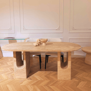 A modern dining room centers on Travertables The Regal Arch Travertine Dining and Meeting Table with distinct curved supports, adorned with a decorative bowl and dried flowers. Its complemented by two black chairs, wooden herringbone flooring, and pristine white walls.
