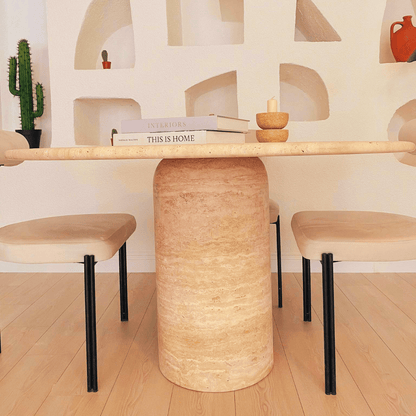 A modern dining area showcases Travertables Chic Oval Travertine Dining Table accompanied by beige cushioned chairs. Books, a candle, and a small bowl adorn the tabletop. A cactus and decorative wall niches enhance the setting, all beautifully laid out on a light wooden floor.