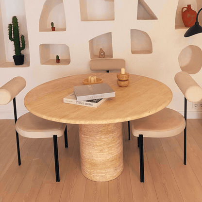The Chic Oval Travertine Dining Table by Travertable, with a central cylindrical base, is encircled by four beige chairs featuring black legs. Books, candles, and bowls adorn the table while recessed white shelves in the background display a small cactus and decor items.