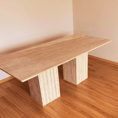 The Ridge Rectangular Travertine Dining Table by Travertable exudes modern sophistication with its sleek top and two wide, fluted pedestal legs, standing gracefully on a hardwood floor against a plain light-colored wall.