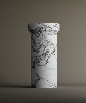 The TRAVERTABLE Calacatta Marble Pedestal Vanity Sink, featuring a tall cylindrical shape with a flared top and striking dark gray veining on a white marble-like base, stands against a plain brown background.